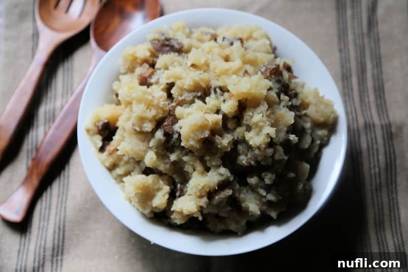 A large white serving bowl filled with creamy Crock Pot Garlic Mashed Potatoes, accompanied by two rustic wooden spoons on a tan placemat.