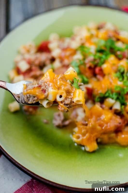 A fork holding a perfect bite of cheeseburger pasta, showcasing the rich cheese and ground beef, ready to be enjoyed from a green plate.