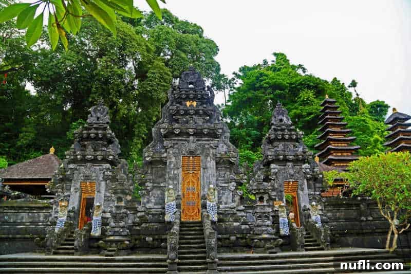 Detailed view of a Balinese temple structure featuring traditional tiered roofs and ornate statues at its entrance, flanked by vibrant green foliage.