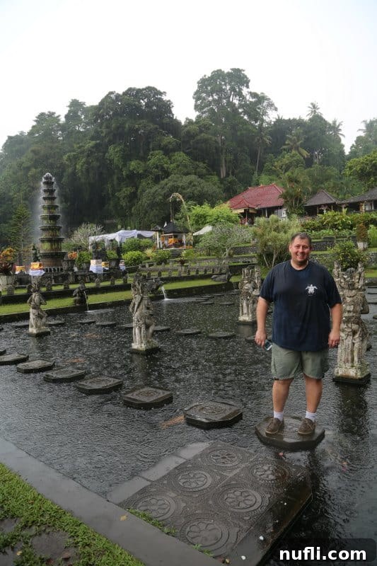 John standing confidently on concrete pavers, elegantly arranged in the middle of a tranquil waterway at Tirta Gangga, surrounded by lush scenery.