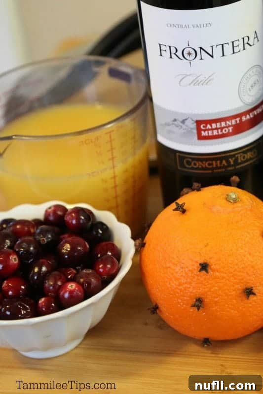 Measuring cup with orange juice, a bottle of red wine, a bowl of fresh cranberries, and an orange studded with whole cloves, ready for mulled wine preparation.