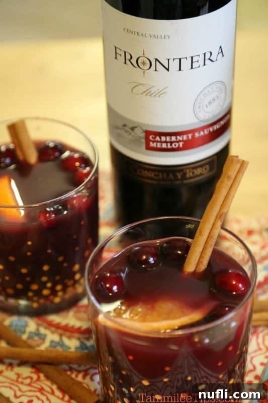 A bottle of red wine stands next to two glasses filled with mulled wine, garnished with fresh cranberries, orange slices, and cinnamon sticks.