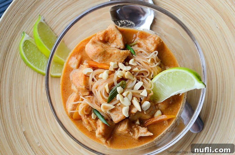 A glass bowl of Thai chicken stew with a lime slice, a spoon, and fresh limes on a wooden board.