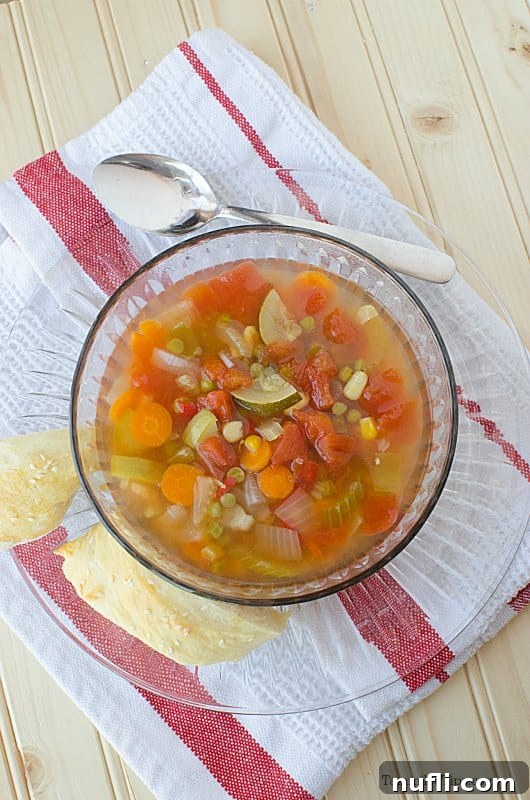 Vegetable soup in a glass bowl sitting on a glass plate next to a silver spoon and cloth napkin, highlighting its rustic appeal