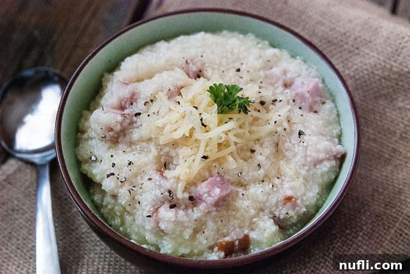 A bowl of creamy ham and cheese grits, perfectly cooked and garnished, with a silver spoon resting on the side, ready for a comforting meal.