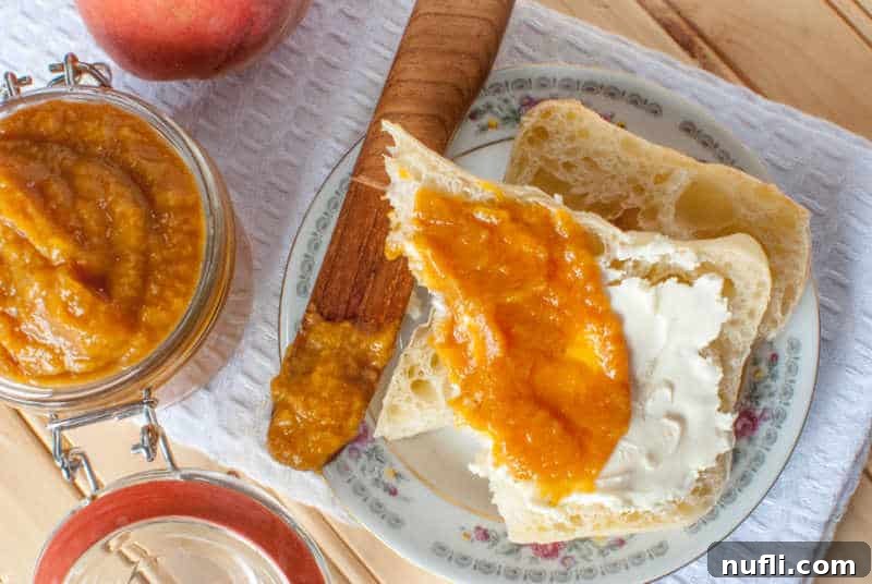 Luscious Crockpot Peach Butter 3 A wooden knife resting on a white napkin next to a fresh peach, a muffin spread with peach butter, and a glass lidded jar filled with Crockpot Peach Butter.