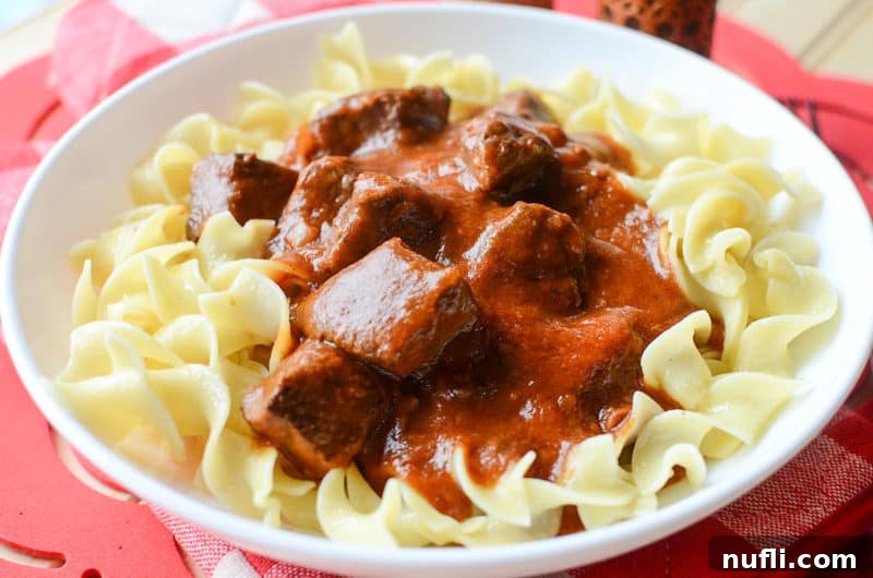 Set It and Forget It Goulash 5 A generous serving of goulash beef over a bed of tender egg noodles on a pristine white plate, invitingly ready to be enjoyed.
