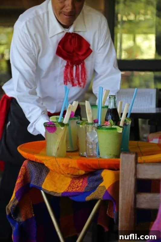 A waiter carrying a tray with two refreshing green beverages, likely margaritas, at an outdoor restaurant