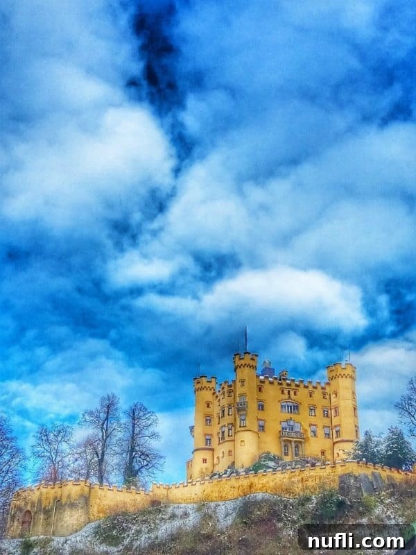Hohenschwangau Castle stands majestically under blue skies with fluffy clouds, overlooking the Bavarian landscape.