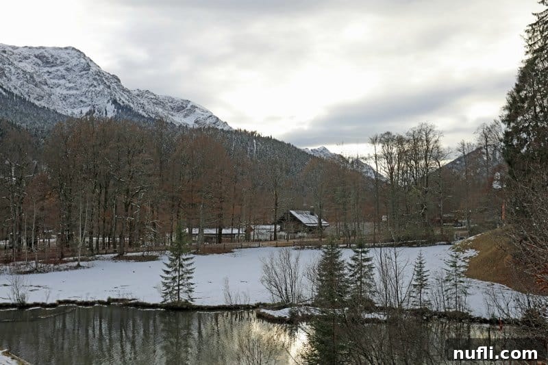 A serene snowy field borders a tranquil pond, with majestic snow-covered mountains rising in the background.