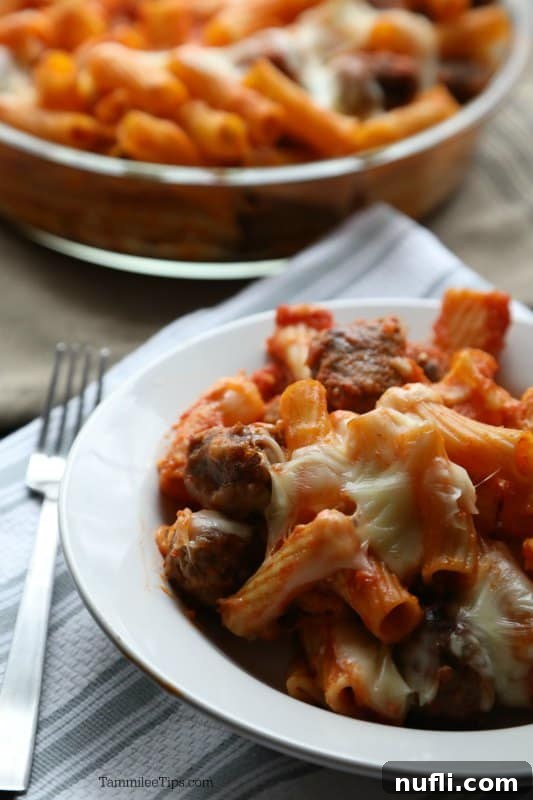 A single serving of baked rigatoni with mini meatballs in a white bowl, next to a fork, showcasing the melted cheese and rich sauce.