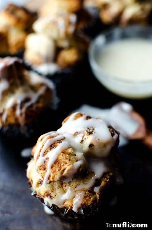 A single maple monkey bread bite, perfectly golden and covered in a sweet white glaze, rests on a dark serving platter. A small bowl of extra icing is visible in the soft-focus background.