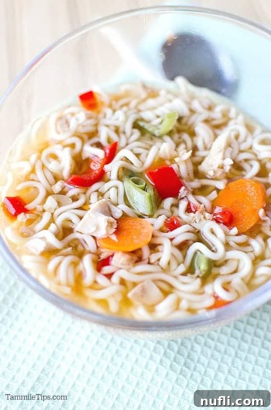 Close-up of a glass bowl filled with rich chicken ramen soup, featuring tender chicken, vibrant vegetables, and noodles, placed next to a spoon on a textured cloth.