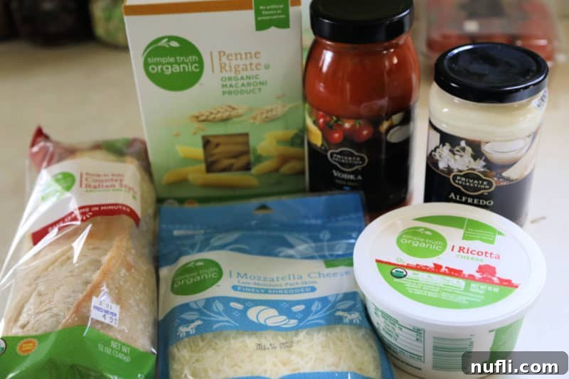 Various ingredients for baked ziti, including dried penne pasta, jars of red sauce and white sauce, and a container of ricotta cheese, neatly arranged on a kitchen counter, ready for preparation.
