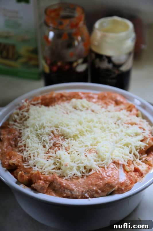 An unbaked baked ziti in a white casserole dish, showing layers of pasta, sauce, and cheese, with empty jars of sauce next to it, indicating preparation in progress.