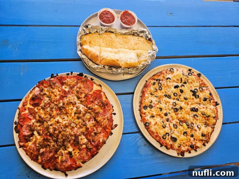 Orange Beach and Gulf Shores Must Try Restaurants 22 two pizzas next to garlic cheese bread with marinara sauce on a blue picnic table