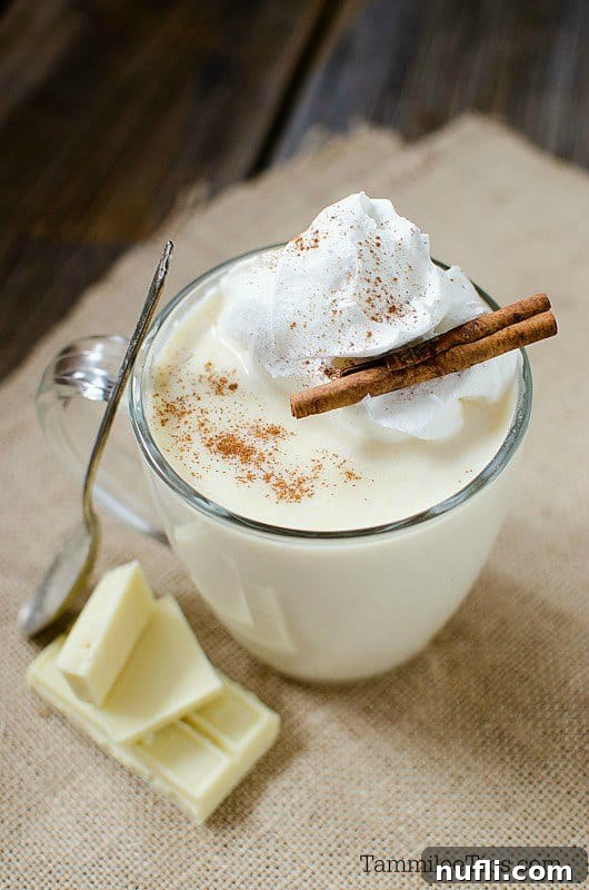 Whipped cream and cinnamon sticks garnishing a white hot chocolate in a glass mug next to a spoon, enhancing its inviting appeal.