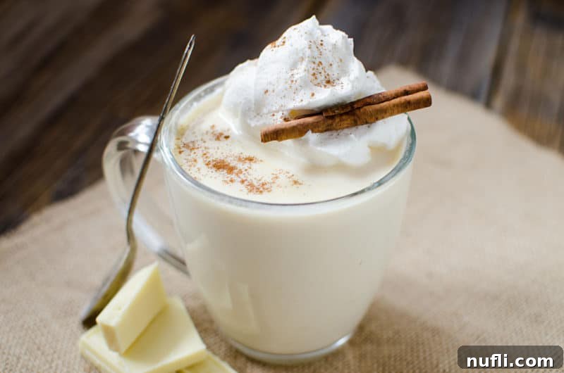 White hot chocolate in a glass mug garnished with whipped cream and a cinnamon stick, invitingly warm.