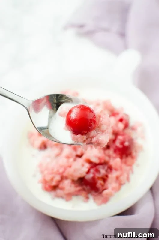 Close-up of a spoon lifting a spoonful of cherry oatmeal, with a whole cherry resting on top, above a bowl of creamy oatmeal.