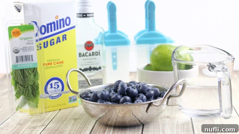 Ingredients laid out for Boozy Blueberry Mojito Popsicles: fresh mint, sugar, Bacardi rum, lime juice, blueberries, and water next to popsicle molds.
