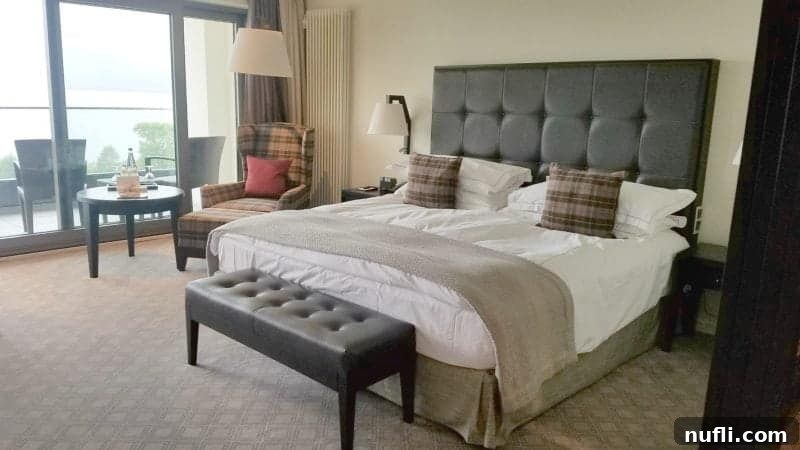 Lakeside Elegance in Killarney: The Europe Hotel 3 Luxurious king bed with a plaid armchair and expansive windows offering stunning lake views at The Europe Hotel Killarney.
