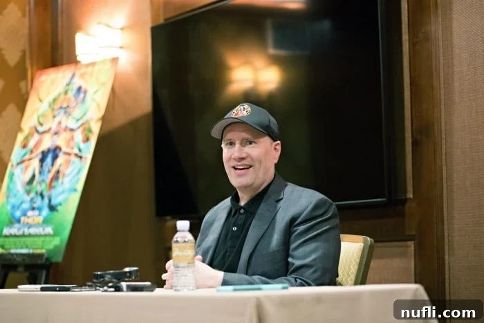 Marvel Studios President Kevin Feige smiles during an interview, seated near a Thor Ragnarok poster.