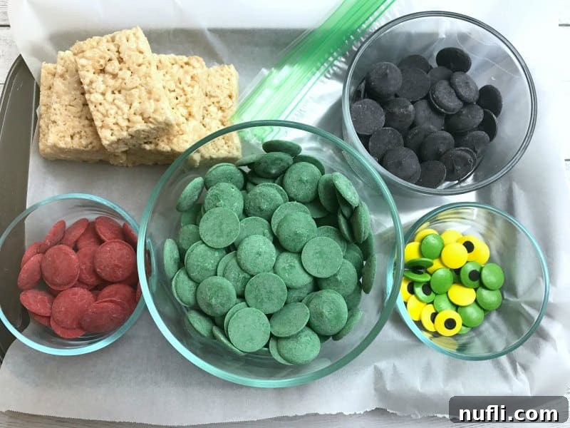 Bowls of red, green, and black candy melts next to a bowl of candy eyes and Rice Krispie treat squares, ready for decoration
