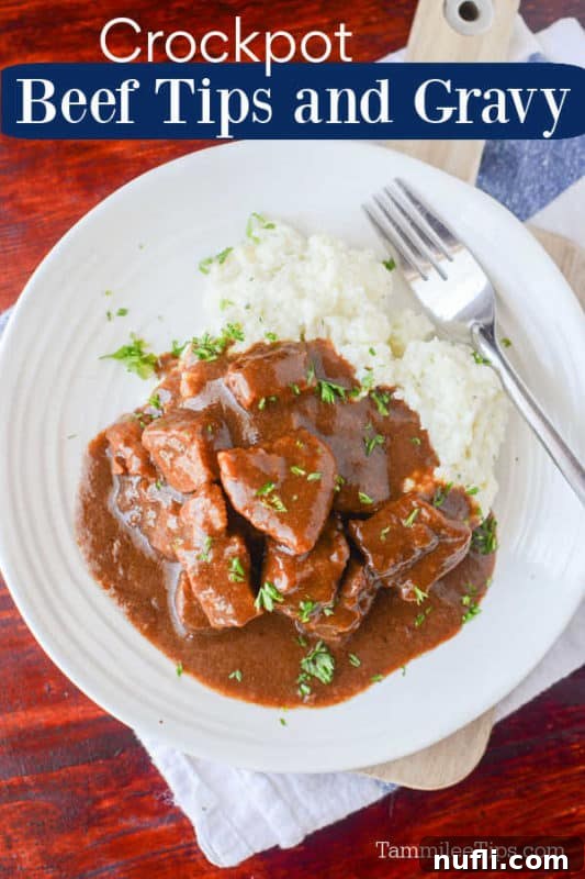 Crockpot beef tips covered in gravy next to mashed potatoes on a white plate with a silver fork on a wood table, emphasizing comfort food