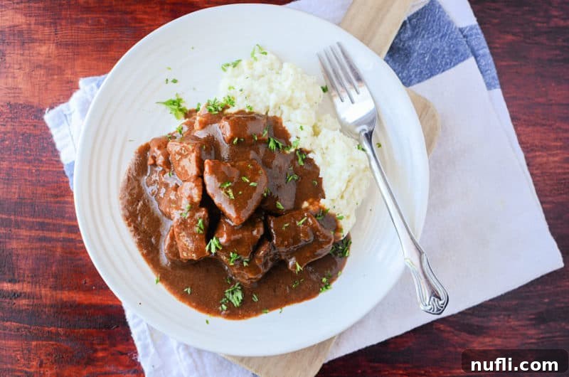 Crockpot beef tips covered in gravy with mashed potatoes on a white plate with a silver fork and cloth napkin, ready for a meal