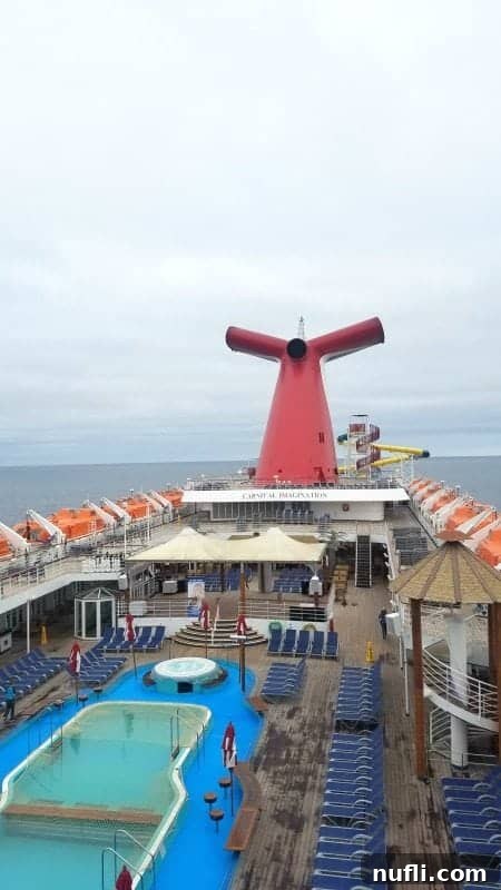 Upper deck of the Carnival Imagination featuring a large outdoor pool surrounded by lounge chairs, under a clear sky.