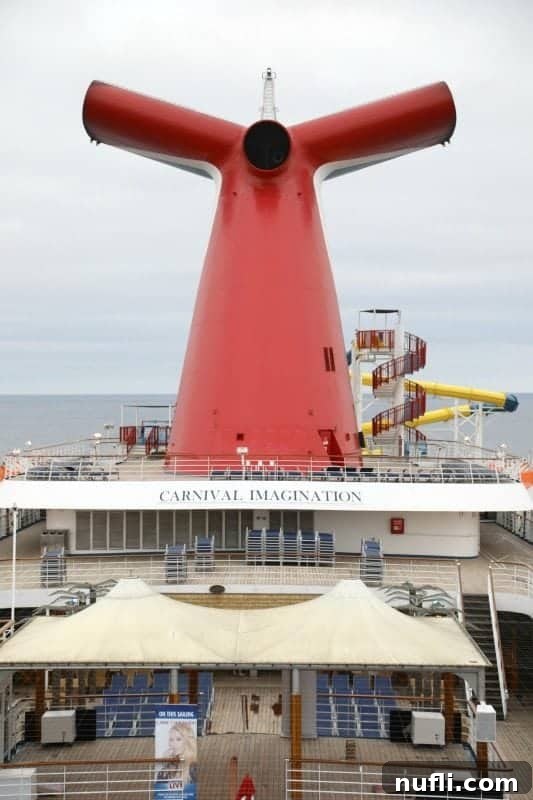 A view of the Carnival Imagination's iconic red, white, and blue funnel against a blue sky, with parts of the upper deck and railings visible.