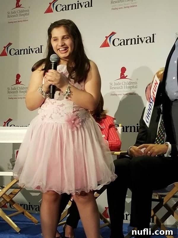 Victoria, a young patient from St. Jude, in a charming pink dress, smiling brightly in front of a Carnival and St. Jude branded backdrop