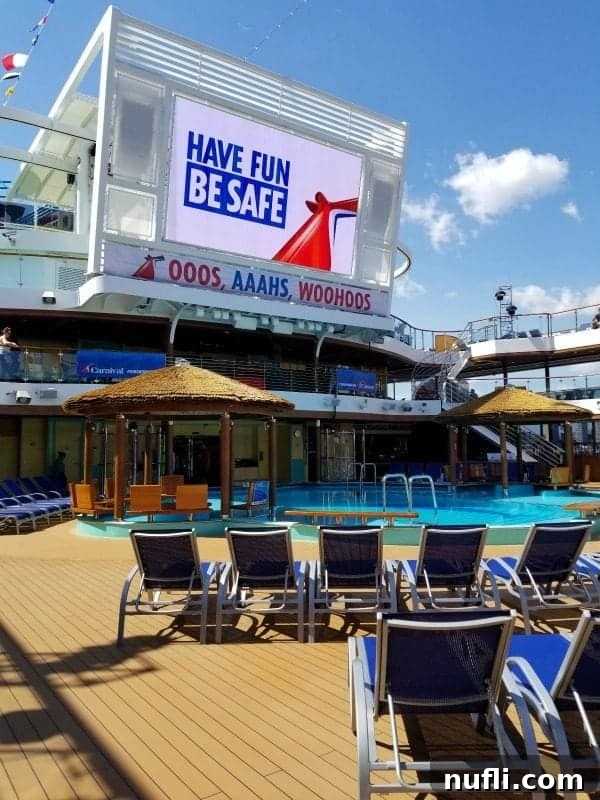 Another perspective of the Carnival Horizon pool deck, featuring a tiki hut, the big TV screen, and numerous blue pool chairs for guests.