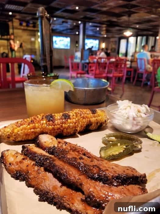 Smoky Bacon Manhattan 6 Platter with strips of beef brisket, corn on the cob, pickled jalapeños, and a cocktail in a restaurant with red chairs and TVs showing sports.
