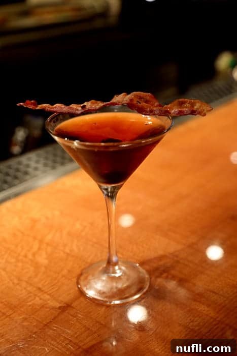 Smoky Bacon Manhattan 9 A single strip of crispy bacon draped over the rim of a cocktail in a martini glass, set against a wooden counter, highlighting the unique garnish.