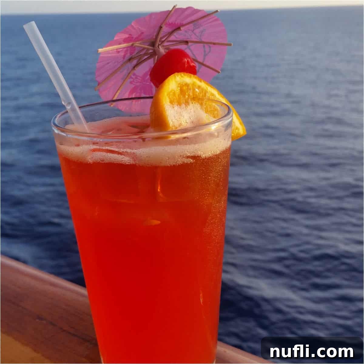 red cocktail with an orange wheel, cherry and pink umbrella with the ocean behind it