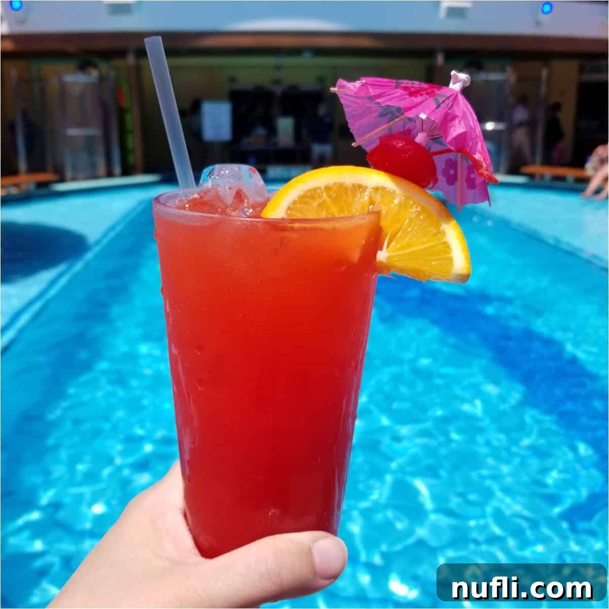 hand holding a red cocktail with orange wheel and pink umbrella by the pool
