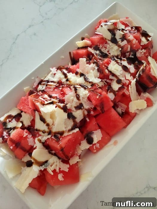 Watermelon salad featuring cubed watermelon, shaved Pecorino Romano cheese, and a rich balsamic glaze on a white platter set against a marble background.