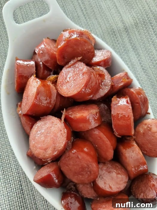 Close-up of Crockpot Brown Sugar Kielbasa bites with a glossy, sweet glaze in a white serving bowl, garnished lightly