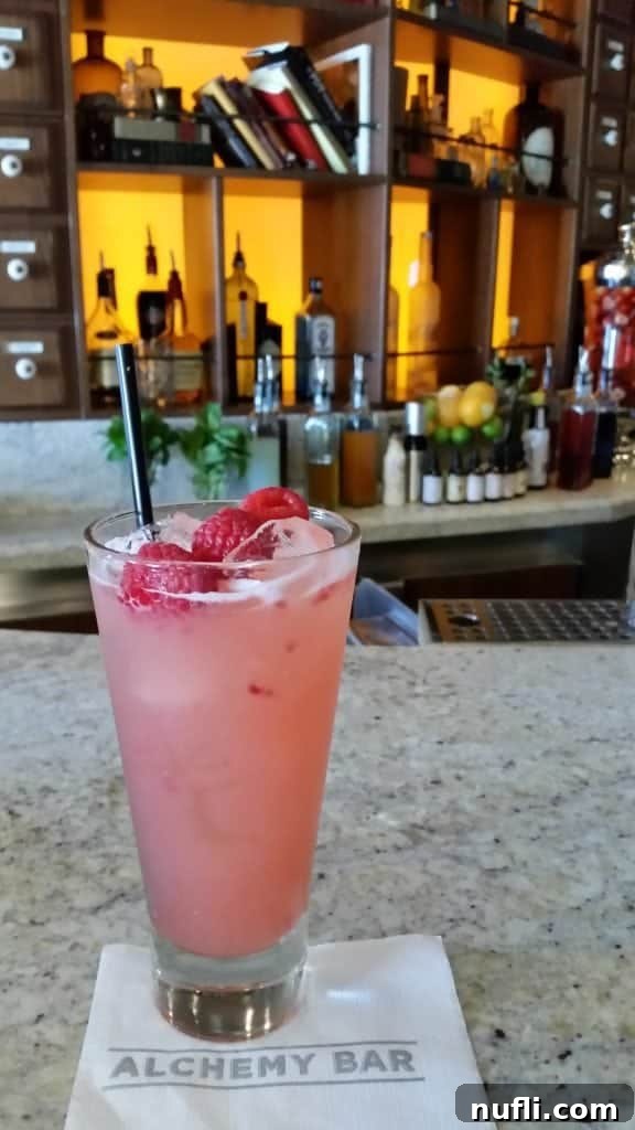 Everything You Need to Know About the Carnival Cheers Package 6 Pink drink with raspberries on a Alchemy bar napkin wtih the bar set up behind it.