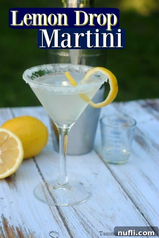 Lemon Drop Martini in a clear glass martini glass with a lemon twist and sugared rim. Cocktail shaker and a couple of lemons behind the cocktail with a white and green background.