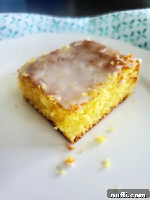 Zesty Lemon Jello Cake Recipe 4 Lemon jello cake square on a white plate next to a cloth napkin