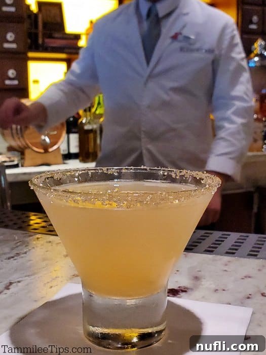 Alchemist Sidecar in a short martini glass, with a skilled bartender in the background behind a well-stocked bar counter.