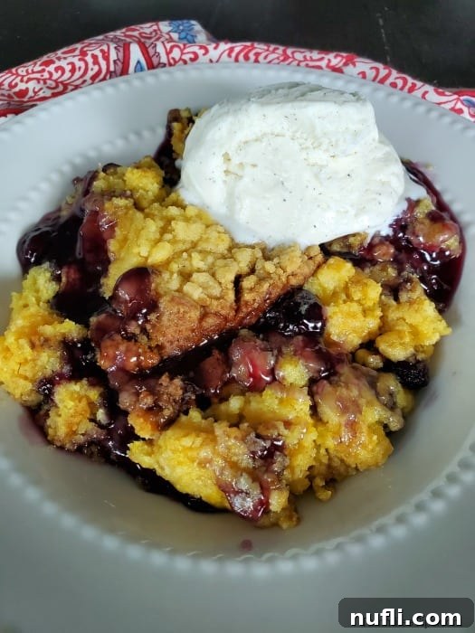 A serving of Crockpot Lemon Blueberry Dump Cake in a white bowl, topped with a scoop of melting vanilla ice cream and fresh blueberries