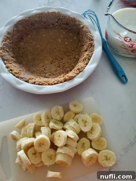 A Nutter Butter crust in a pie dish, accompanied by a whisk, measuring cup, and neatly sliced bananas on a cutting board, ready for assembly.