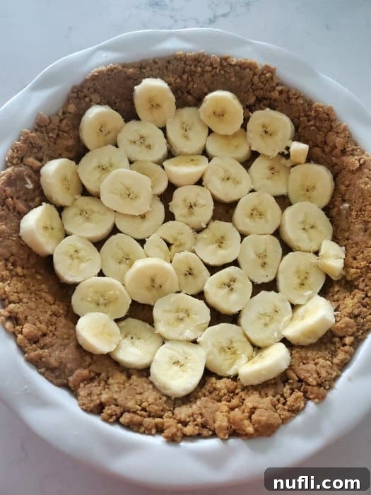 Neatly sliced bananas laid over the Nutter Butter crust in a white pie plate, forming the first layer of the pudding.