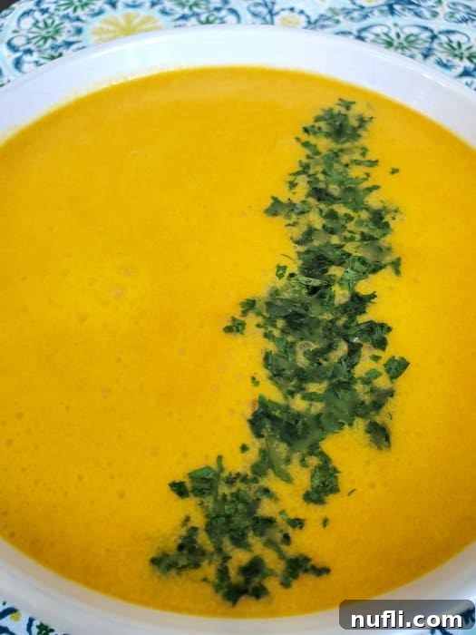A serving of vibrant orange carrot ginger soup, garnished with a green leafy herb, presented in a white bowl on a colorful patterned napkin