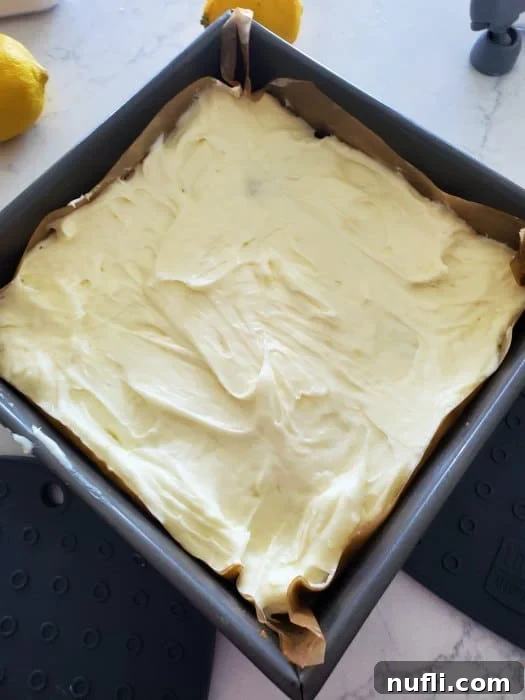 Smooth lemon cheesecake filling spread evenly over the pre-baked crust in a parchment-lined baking sheet, topped with a final crumble layer.