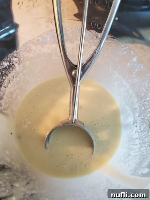 The 3 Ingredient Muffin Mix Pancake Hack 4 A medium-sized cookie scoop submerged in a bowl of creamy, light-colored muffin mix pancake batter, ready to be cooked.