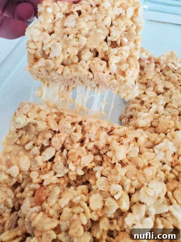 Delicious Peanut Butter Rice Krispie Treats 3 A hand reaching into a glass baking dish to pick up a square of freshly made peanut butter rice krispie treat, showcasing its texture and portion size.